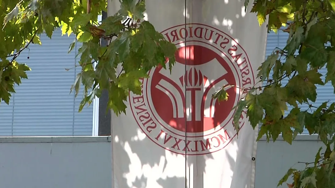 Bijela zastava sa crvenim grbom univerziteta i natpisom na latinskom, djelimično zaklonjena granama drveta sa zelenim lišćem.