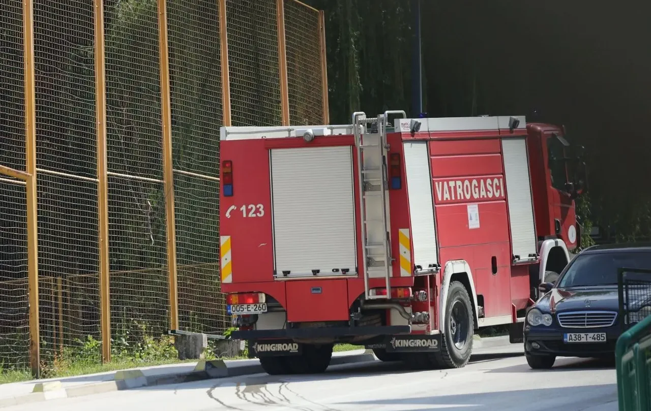 Crveno vatrogasno vozilo sa natpisom "Vatrogasci" i brojem "123", parkirano pored tamnog automobila, sa žutom ogradom u pozadini.