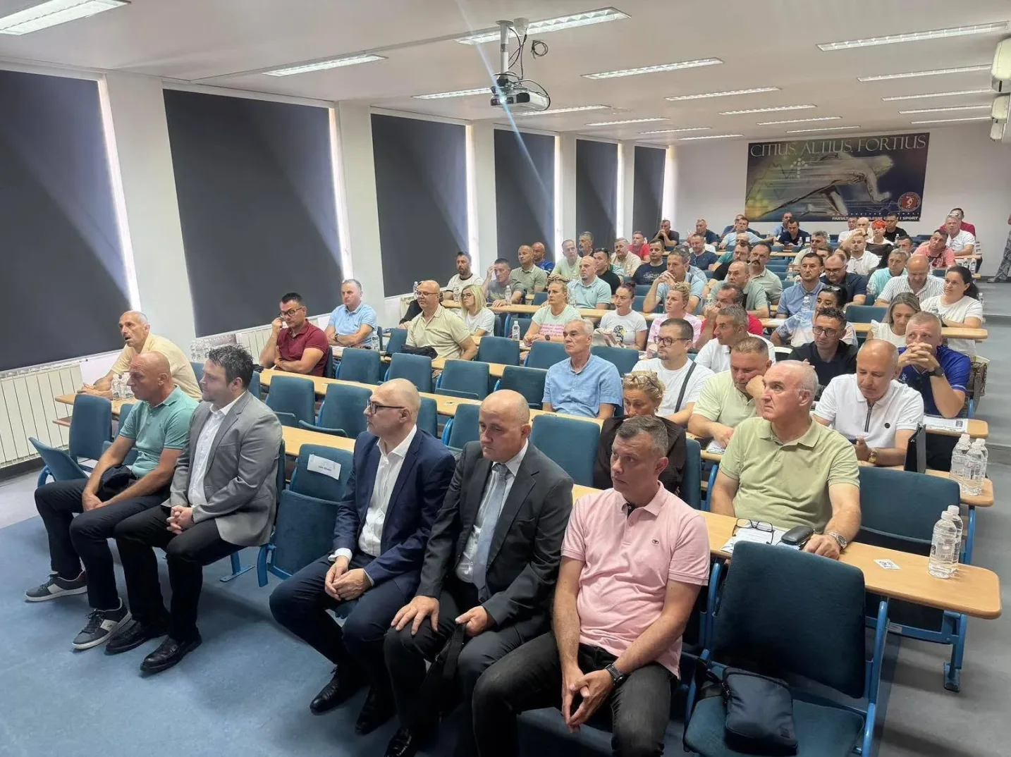 Grupa ljudi sjedi u predavaonici, gledaju u smjeru predavača.