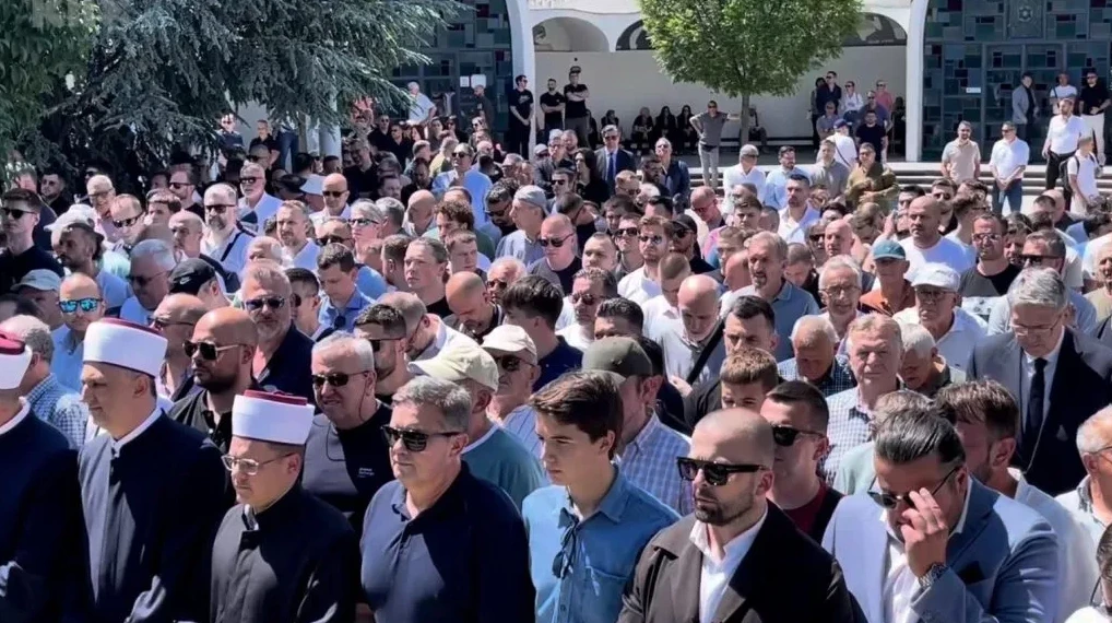 Velika grupa ljudi, uglavnom muškaraca, okupljena vani na događaju, vjerovatno vjerskom ili zvaničnom. U prvom planu se ističu neki muškarci sa fesovima, sugerišući vjerski karakter skupa. Mnogi od prisutnih nose sunčane naočale. U pozadini se naziru zgrada i drveće.