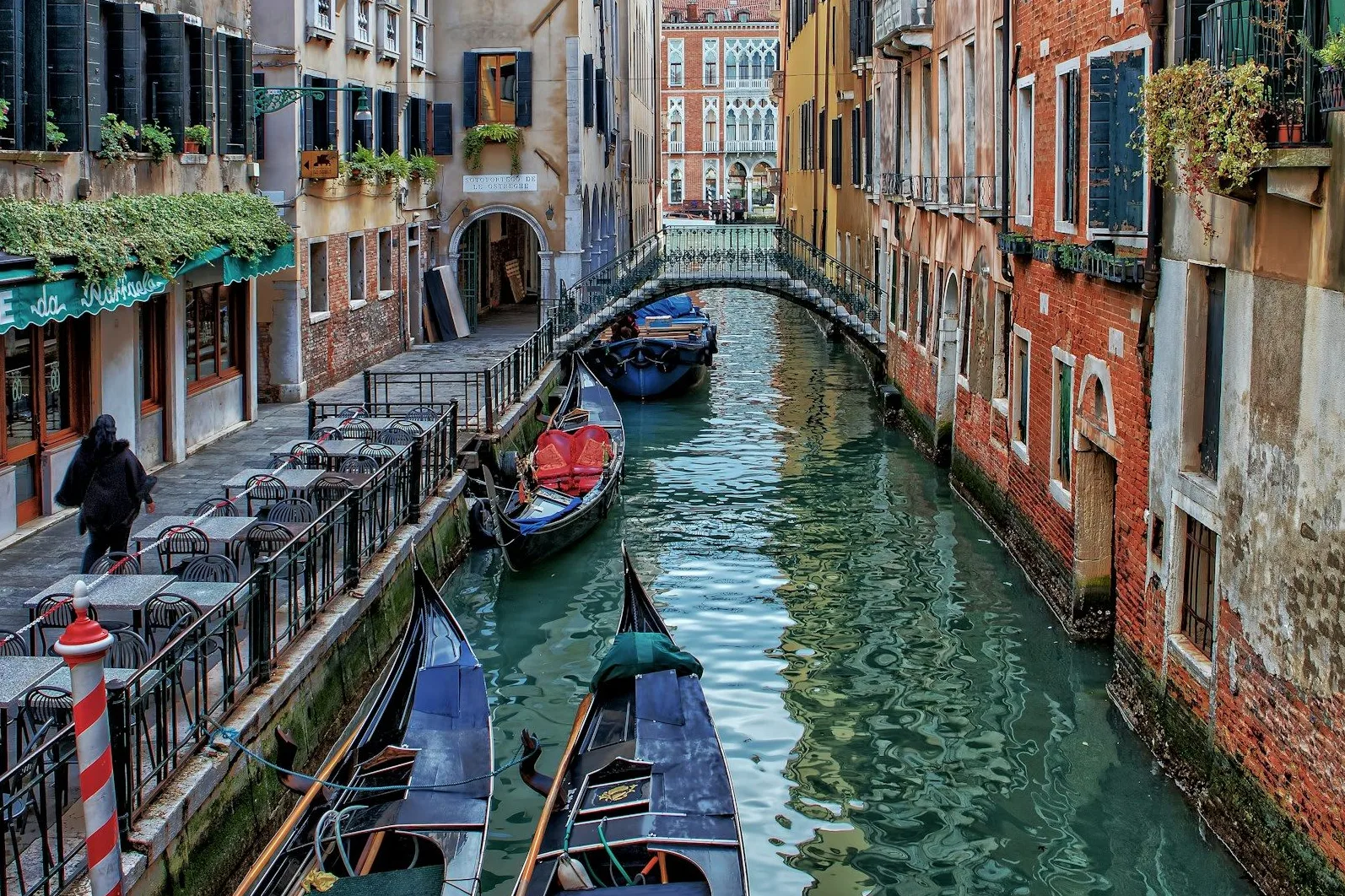 Pogled na uski venecijanski kanal okružen starim zgradama od cigle i maltera, sa nekoliko gondola usidrenih uz obalu i pontom sa stolovima i stolicama.