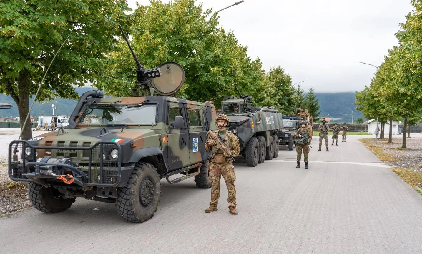 Italijanski vojnici u punoj opremi stoje pored oklopnog vozila naoružanog mitraljezom i drugih vojnih vozila u formaciji.