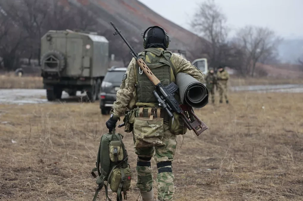 Vojnik u vojnoj uniformi sa snajperskom puškom na leđima i ruksakom u ruci.