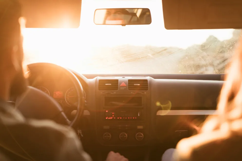 Pogled iz unutrašnjosti automobila prema naprijed, vozač sa lijeve strane, suvozač sa desne, sa suncem koje sija kroz vjetrobransko staklo.