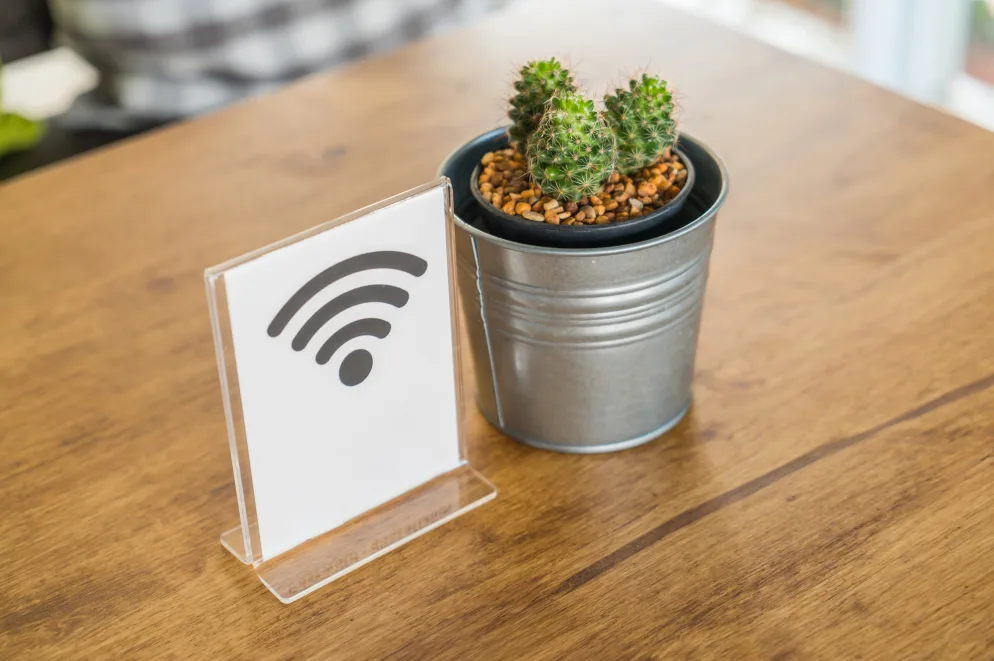 Bijeli znak sa simbolom za bežični internet (Wi-Fi) i malim kaktusom u metalnoj saksiji na drvenom stolu.