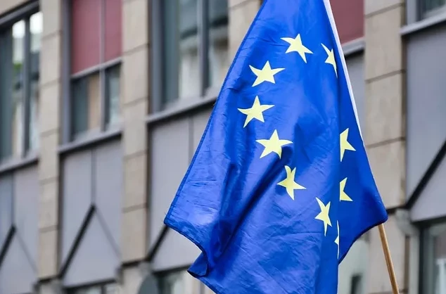 Zastava Evropske unije (EU) sa zlatnim zvjezdicama na plavoj pozadini, u pokretu, ispred moderne zgrade sa staklenim prozorima.