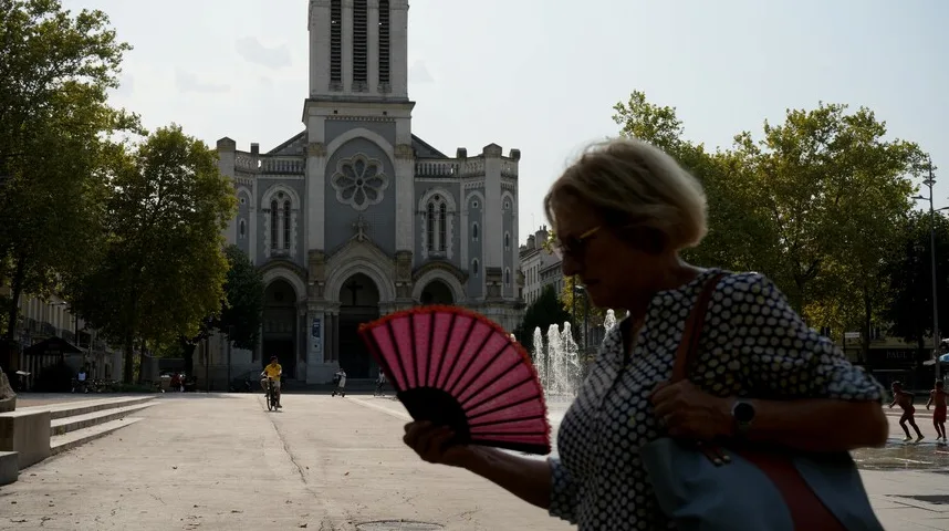 Žena u prvom planu drži rasklopljenu crvenu lepezu ispred sebe, hladeći se po vrućem danu. U pozadini se nazire impozantna crkva sa tornjem i rozetom, okružena drvećem. Na trgu je takođe vidljiv fontana i nekoliko ljudi, uključujući dijete koje se vozi biciklom i djecu koja se igraju u vodi.