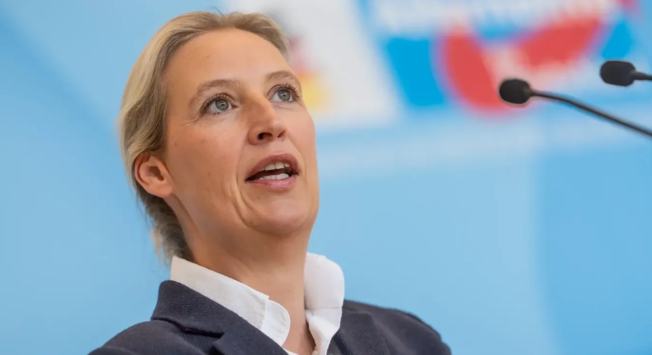 Žena sa plavom kosom govori za govornicom ispred mikrofona i plave pozadine sa logom stranke