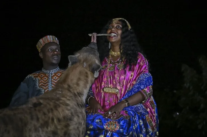 Žena u tradicionalnoj afričkoj nošnji dijeli hranu sa hijenom, dok muškarac u sličnoj odjeći stoji iza nje.