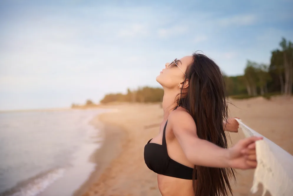 Žena u crnom bikiniju sa raširenim rukama na plaži, gleda prema nebu.