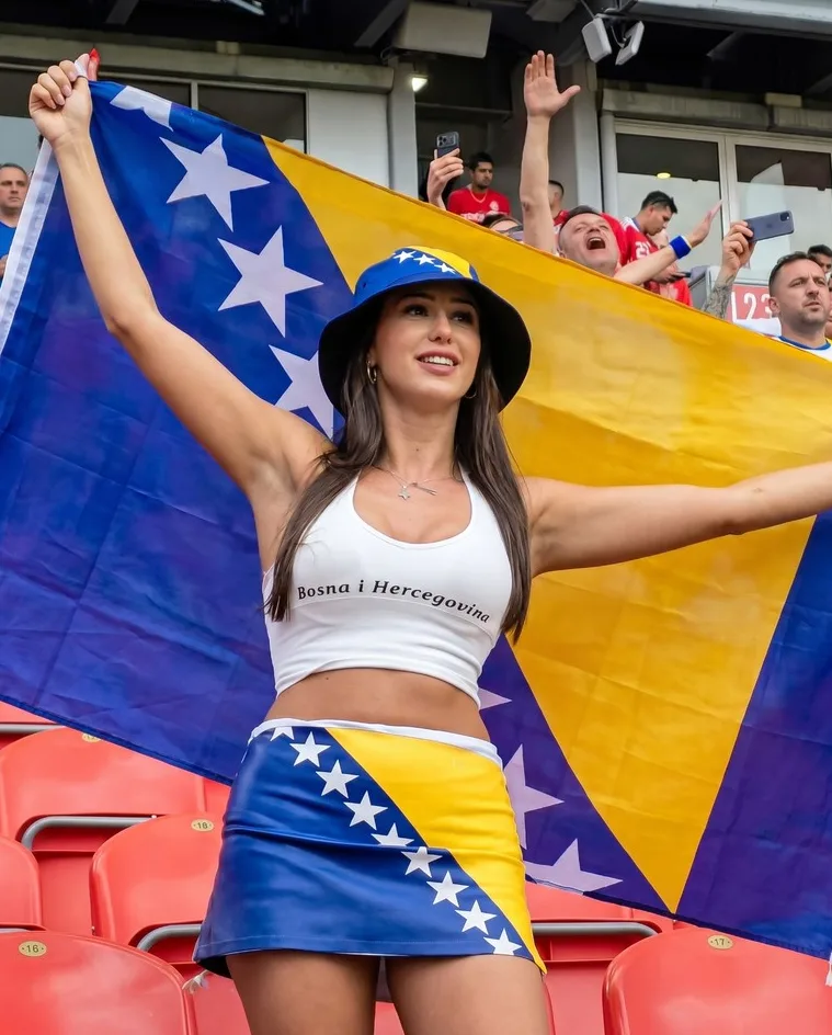 Mlada navijačica Bosne i Hercegovine na stadionu drži zastavu svoje zemlje i nosi šešir te suknju u bojama zastave.