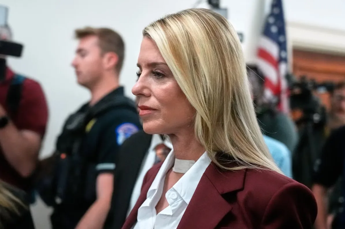 Žena u crvenom svecanom odijelu i bijeloj košulji, sa trakom oko vrata, gleda lijevo u okruženju sa prisutnim osobljem i američkom zastavom u pozadini.
