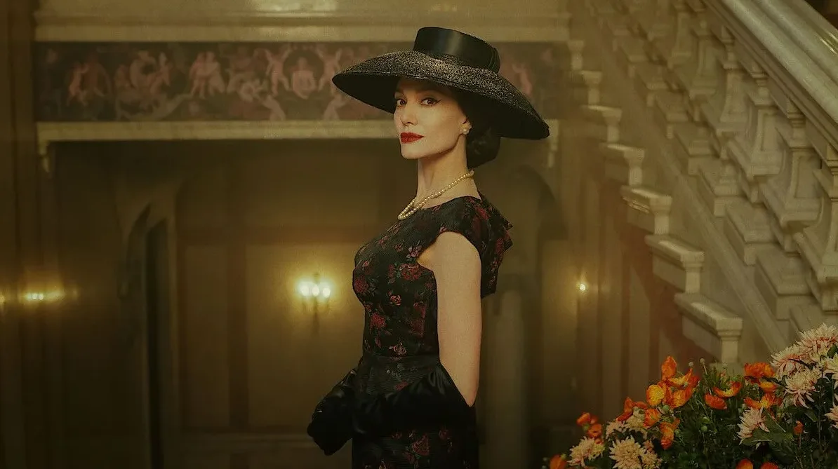 Žena u elegantnom crnom šeširu, bisernoj ogrlici i tamnoj haljini s cvjetnim uzorkom, pozira pored stepeništa.