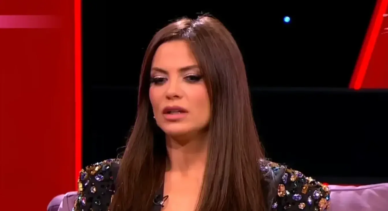 Žena sjedi u studiju s crvenom pozadinom i crnim ekranom u pozadini, noseći ukrašenu haljinu.