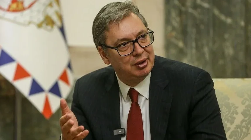 Aleksandar Vučić u odijelu i sa crvenom kravatom nosi naočale i gestikulira desnom rukom, sa zastavom u pozadini.