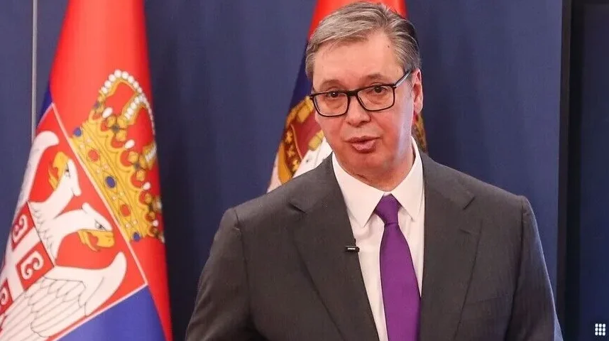 Aleksandar Vučić u sivom odijelu i ljubičastoj kravati, s naočalama, stoji ispred zastava Srbije.