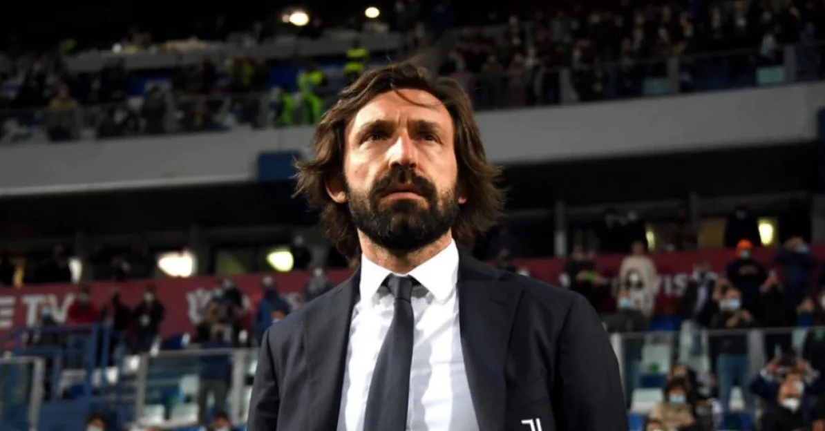 Andrea Pirlo, trener Juventusa, u crnom odijelu i bijeloj košulji s tamnom kravatom, gleda prema naprijed na stadionu.