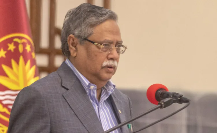 Bangladeški zvaničnik sa Bijelom bradom i naočarima govori za govornicom sa mikrofonom ispred zastave Bangladeša.