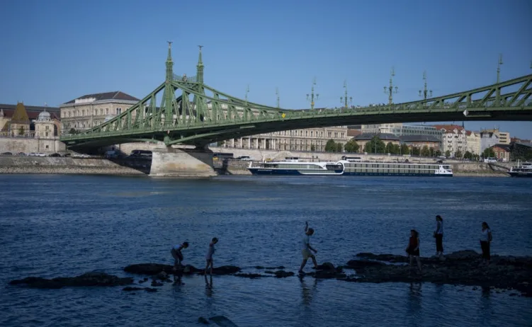 Siluete ljudi na kamenju u Dunavu ispred zelenog mosta i zgrada u Budimpešti.
