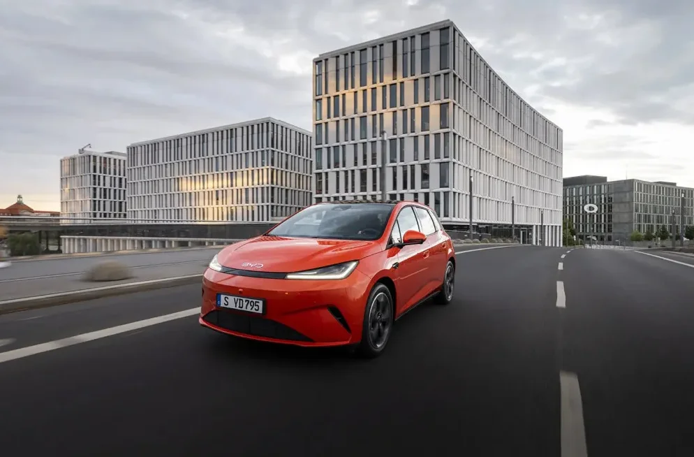 Narandžasti BYD električni automobil u pokretu na putu ispred arhitektonski modernih zgrada.