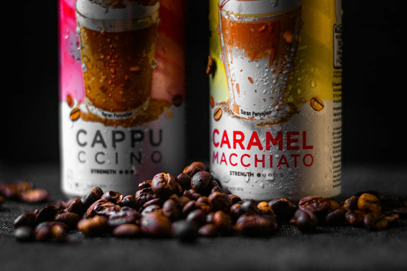 Dvije limenke kafe, jedna "Cappuccino" i druga "Caramel Macchiato", sa kapljicama vode, ispred hrpe prženih zrna kafe na tamnoj pozadini.