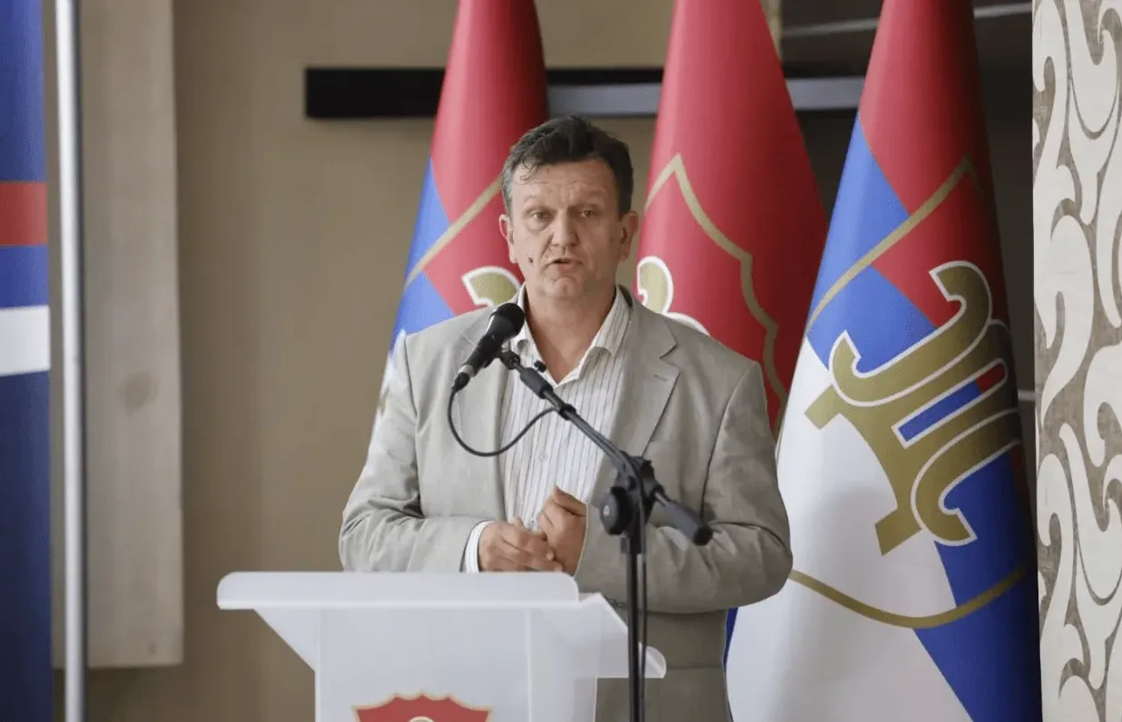 Muškarac u svijetlom sakou govori u mikrofon iza postolja, a iza njega se nalaze zastave.