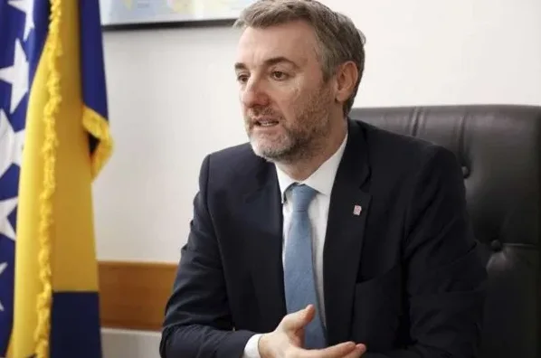 Čovjek u tamnom odijelu i plavoj kravati sjedi u crnoj kožnoj fotelji, s rukama ispruženim ispred sebe. U pozadini se nazire zastava Bosne i Hercegovine.