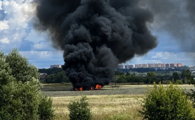 Crni gusti dim suklja iznad zapaljenog polja, s nizom stambenih zgrada u daljini.