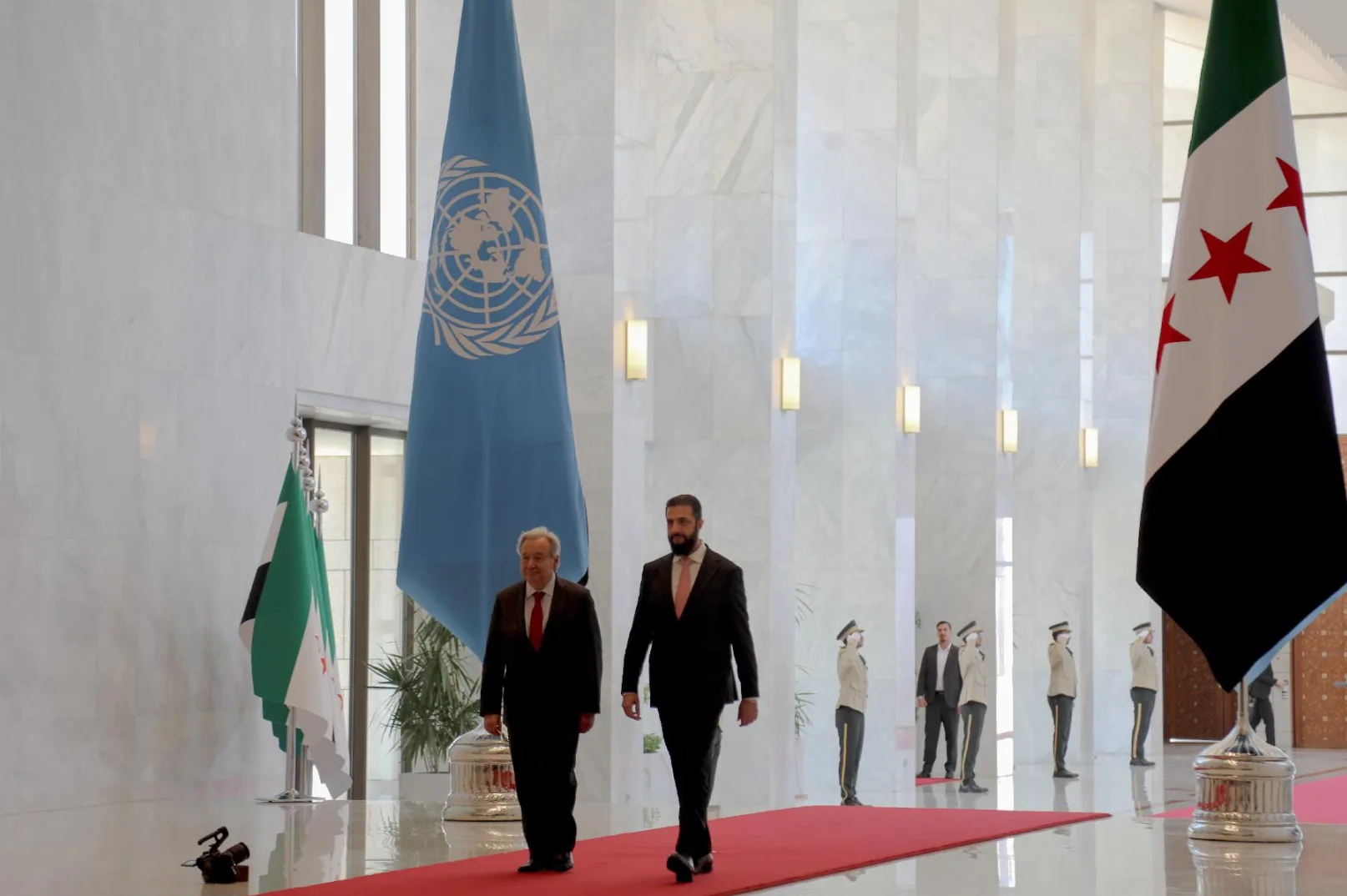 Dva diplomata hodaju crvenim tepihom, sa zastavom Ujedinjenih naroda (UN) lijevo i sirijskom zastavom desno. Muškarac u tamnom odijelu s crvenom kravatom hoda ispred muškarca u tamnom odijelu s ružičastom kravatom. U pozadini se nalaze ljudi u uniformama koji stoje u stavu mirno, te drugi diplomata koji ih promatra.