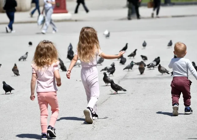 Dvoje male djece i beba šetaju među mnoštvom golubova na gradskom trgu po sunčanom danu.