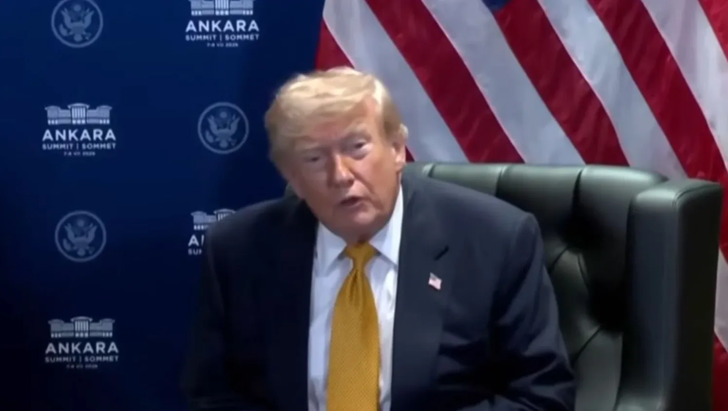 Donald Trump sjedi u crnoj kožnoj stolici, noseći tamno odijelo sa žutom kravatom. U pozadini se vidi američka zastava i plavi panel sa logom Ankarskog samita.
