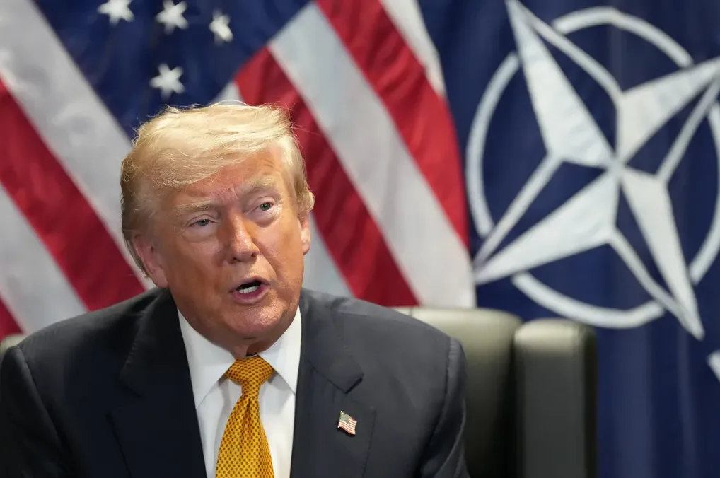 Donald Trump u odijelu, sa žutom kravatom, govori. U pozadini se vide zastave Sjedinjenih Američkih Država i NATO-a.