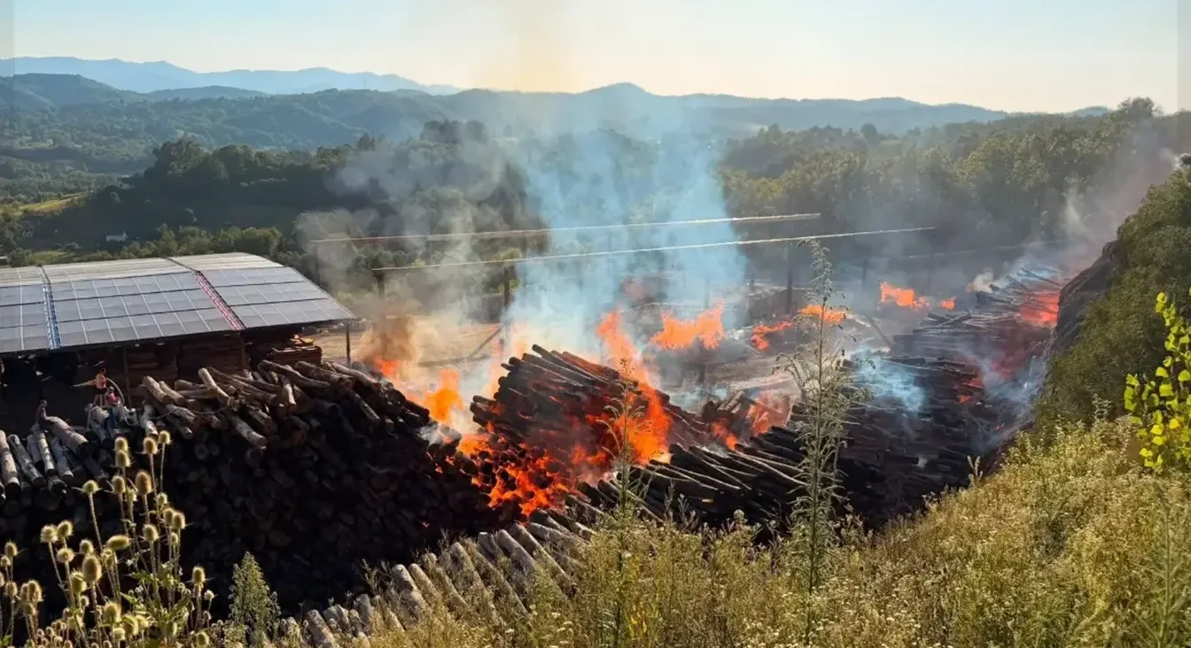 Veliko skladište drvenih trupaca u plamenu, gust dim se diže u vazduh. U pozadini se vide zeleni brežuljci i plavo nebo.