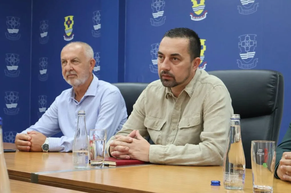 Dva muškarca sjede za stolom, obojica gledaju ulijevo, s vodom i čašama ispred sebe, na pozadini plavog zida s logotipima.