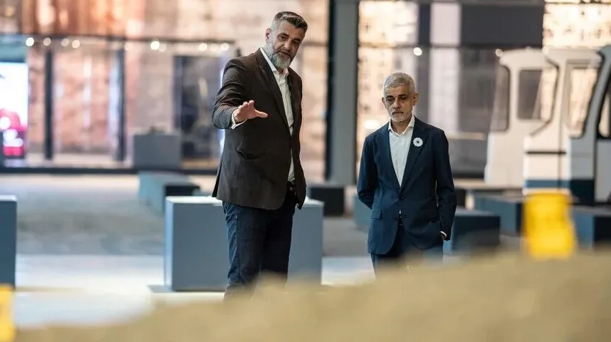 Dva muškarca u odijelima stoje u modernom prostoru, jedan od njih gestikulira rukom.