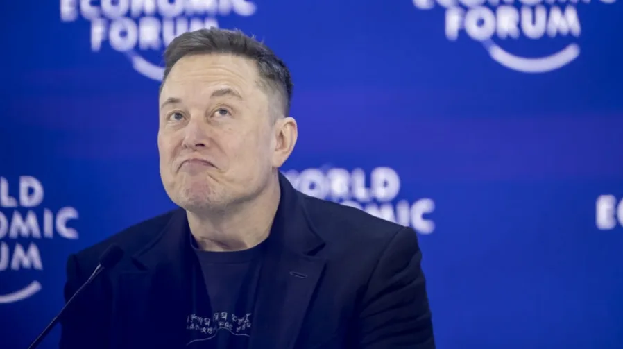 Elon Musk sa smiješkom na licu i podignutim obrvama, u crnom sakou, sa logom Svjetskog ekonomskog foruma u pozadini.
