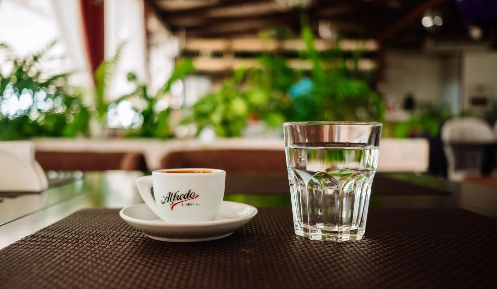 Šoljica espresa s logom 'Alfredo Espresso' pored čaše hladne vode na stolu u restoranu.