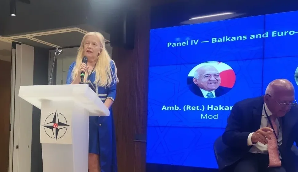 Žena u plavom kostimu drži mikrofon i stoji za govornicom sa logom NATO-a. Iza nje, na ekranu se vidi tekst "Panel IV - Balkans and Euro-" i portret muškarca sa natpisom "Amb. (Ret.) Hakani Mod".