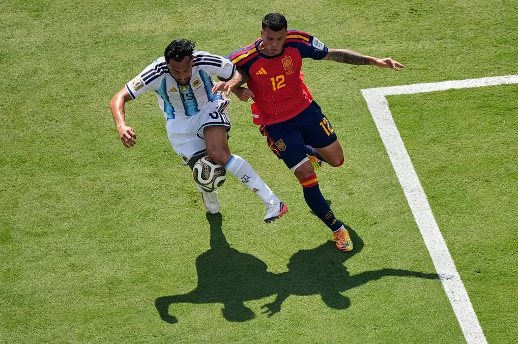 Fudbaler Argentine u bijelo-plavom dresu u duelu sa fudbalerom Španije u crvenom dresu na zelenom terenu.