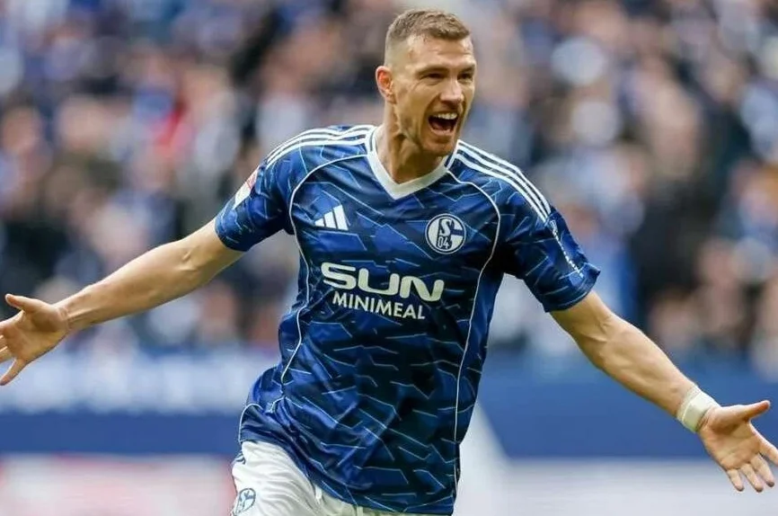 Fudbaler u plavom dresu sa bijelim prugama i grbom kluba Schalke 04, raširenih ruku i uzvikujući od sreće nakon postignutog gola.