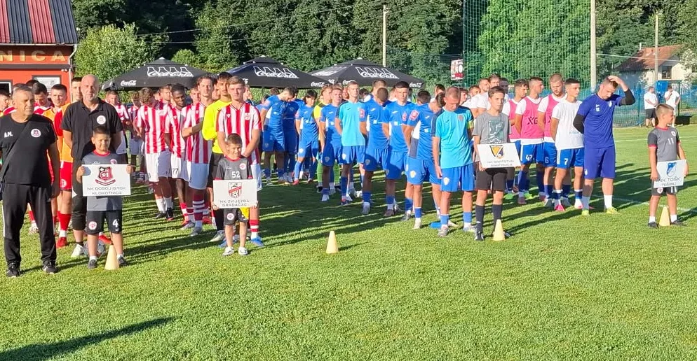 Grupa mladih fudbalera, podijeljenih u dva tima (jedan u plavim dresovima, drugi u crveno-bijelim prugastim dresovima), stoje na zelenom travnjaku fudbalskog igrališta. Neki od dječaka nose transparente sa logom i imenima svojih klubova. U prvom planu vide se treneri ili stariji pratitelji. U pozadini se naziru suncobrani i drveće, a sve ukazuje na sportski događaj ili utakmicu.
