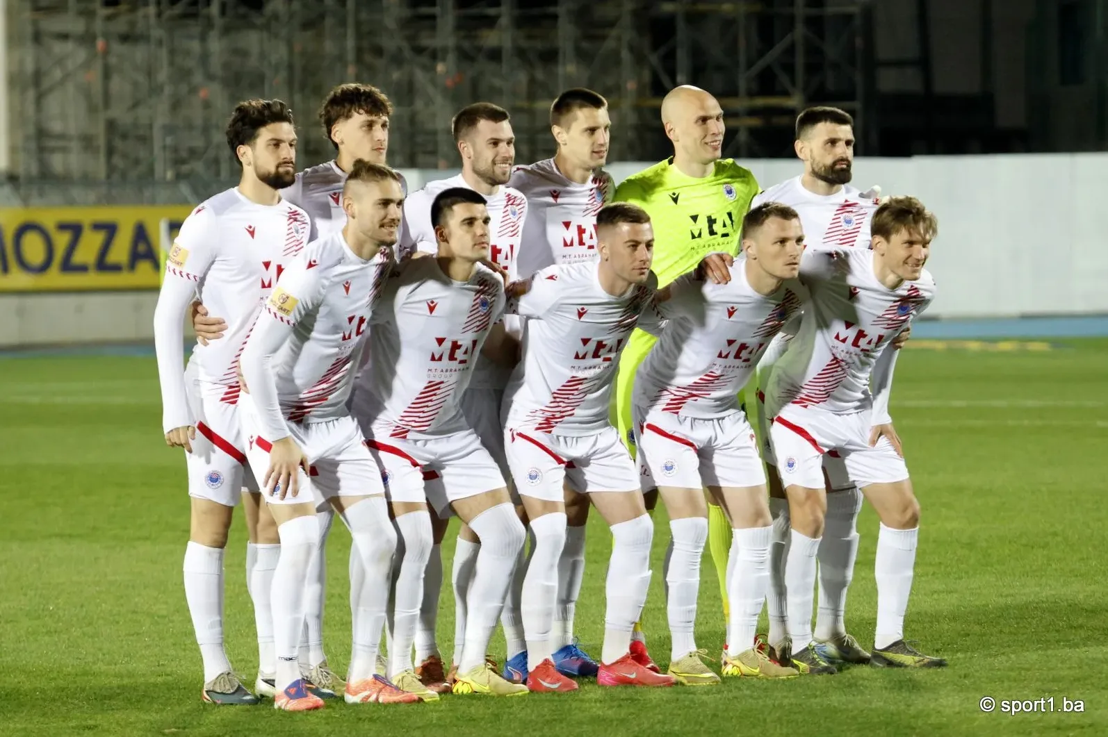 Fudbalski tim u bijelim dresovima sa crvenim detaljima pozira na zelenom terenu.