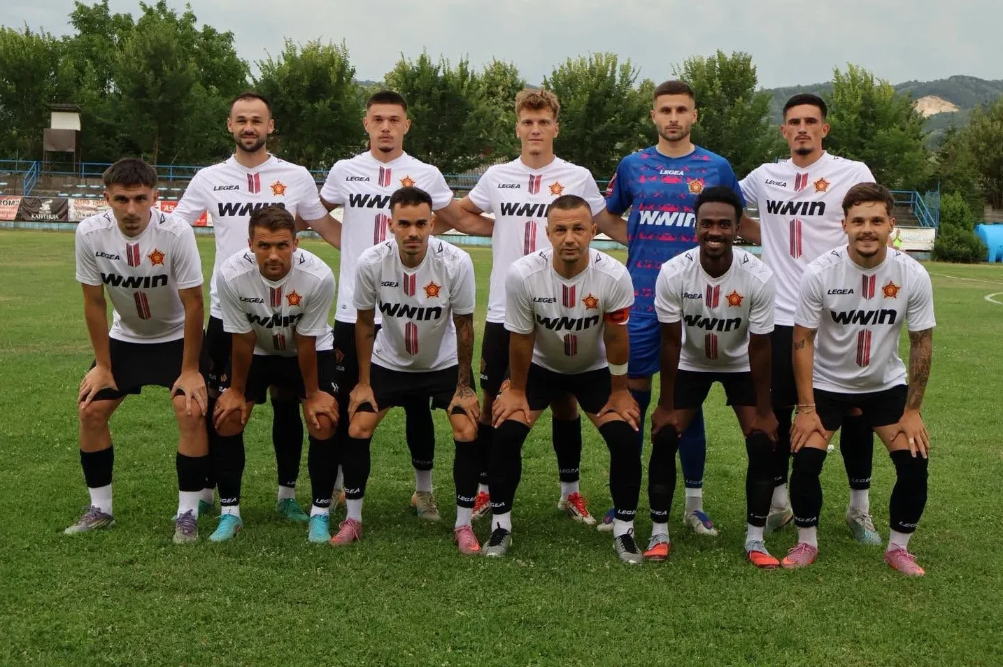 Grupa fudbalera u bijelim dresovima sa crvenim detaljima i crnim šorcićima, pozira na zelenom travnjaku fudbalskog stadiona.