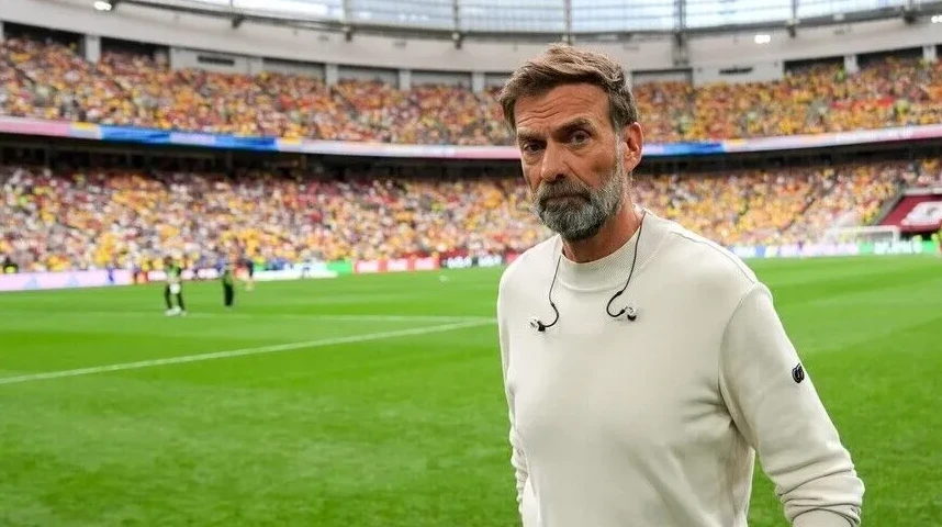 Fudbalski trener sa bradom u bijeloj majici sa slušalicama oko vrata, stoji na zelenom terenu stadiona