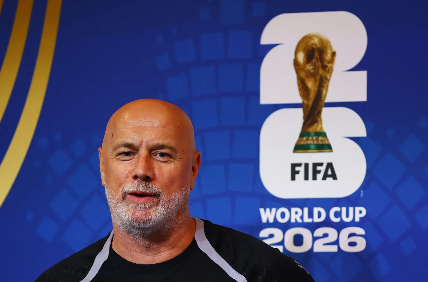 Muškarac kratke sijede kose i brade, obučen u crnu majicu, stoji ispred plave pozadine sa logotipom FIFA Svjetskog kupa 2026.