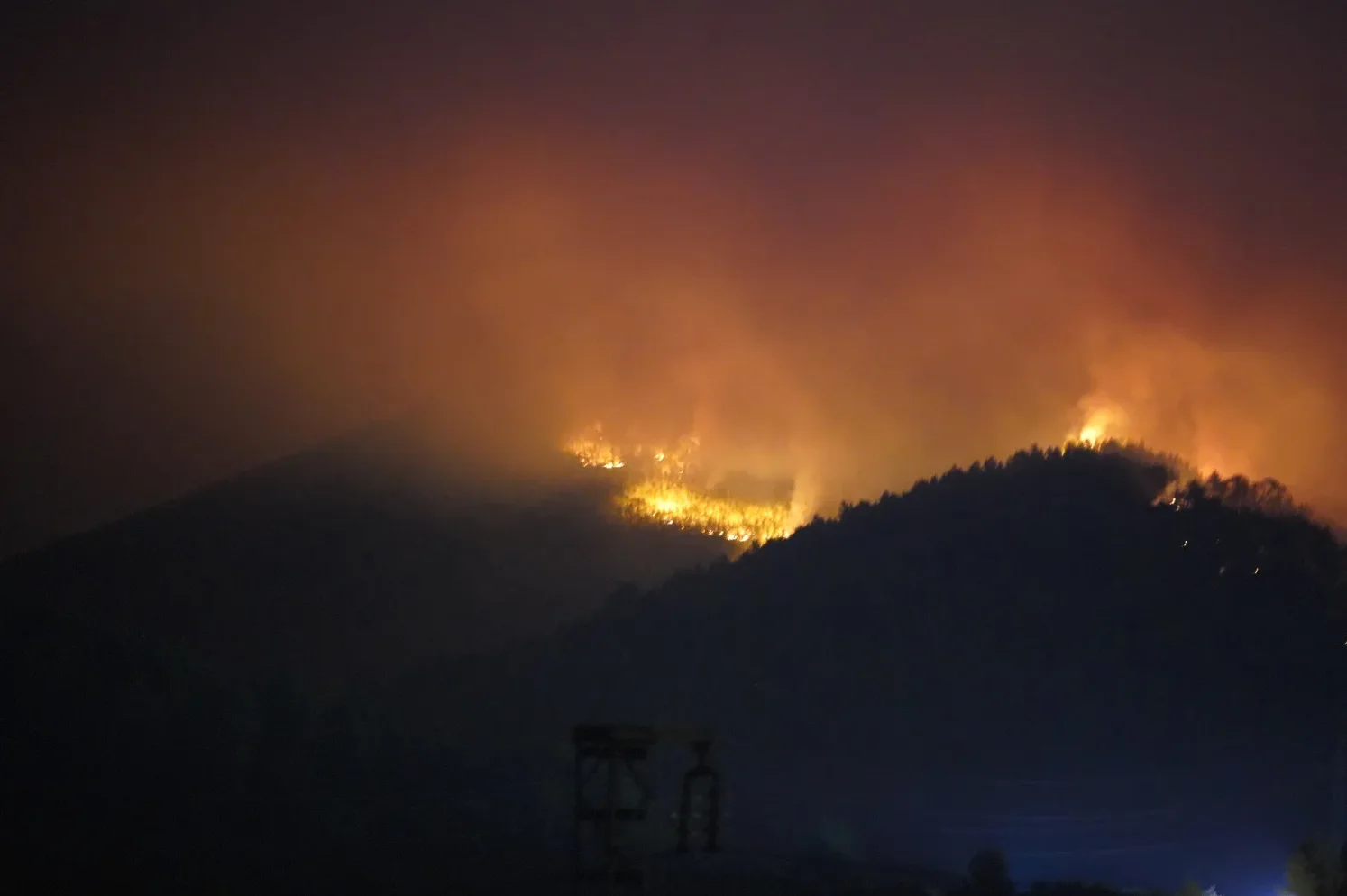 Žuti plamenovi gorućeg požara osvjetljavaju tamnu planinsku padinu noću, sa gustim narandžastim dimom koji se diže u nebo.