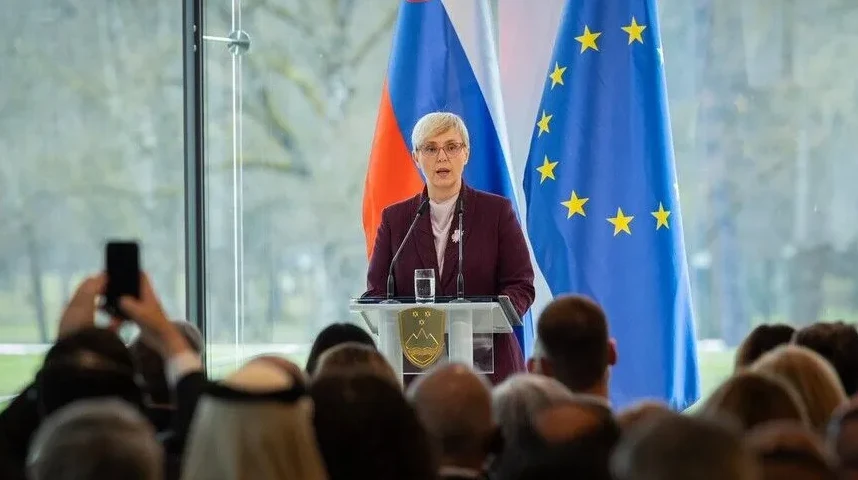 Žena u tamnom sakou i bijeloj košulji, sa kratkom plavom kosom i naočalama, govori za govornicom.