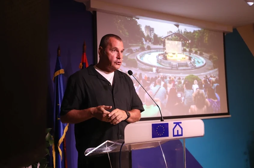 Muškarac u crnoj košulji govori za govornicom ispred projektovanog prizora fontane i publike.