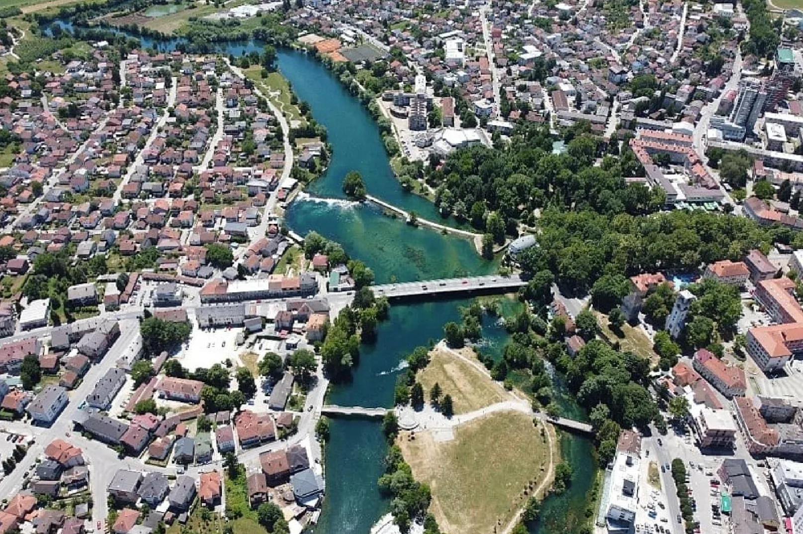 Aerijalni snimak grada sa rijekom koja protiče kroz njega, vidljiva su dva mosta sa saobraćajem, zelene površine i krovovi kuća.