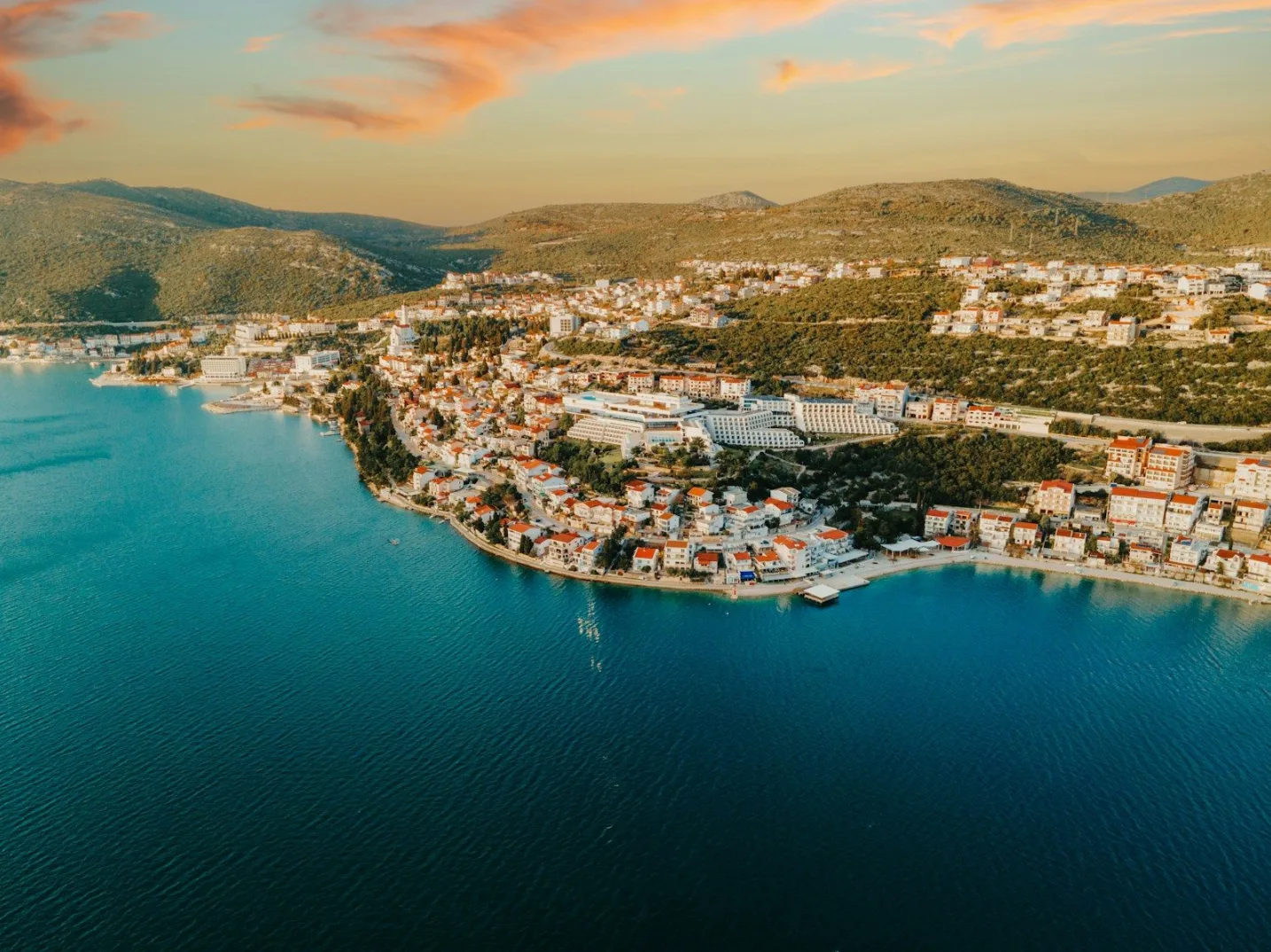 Aerijalni pogled na grad sa bijelim zgradama i crvenim krovovima, smješten na zelenoj padini iznad tirkiznog mora.